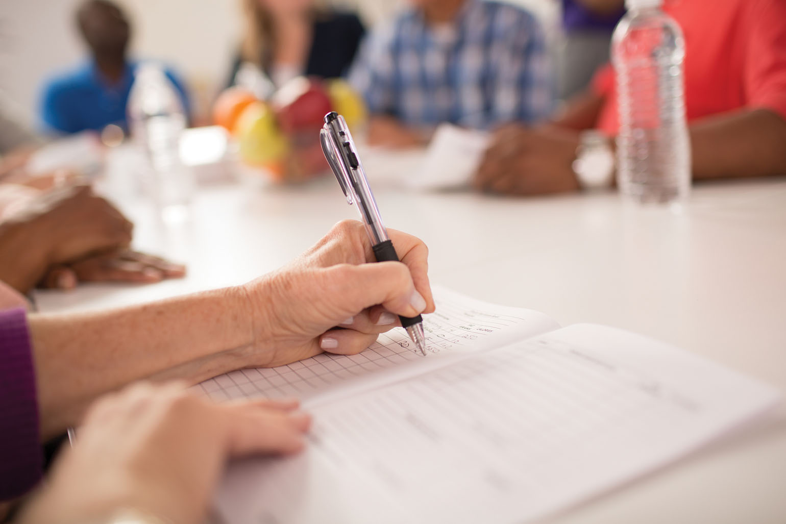 annual-reports A close-up of a person writing notes in a notebook during a group meeting, with blurred participants and fruit in the background.