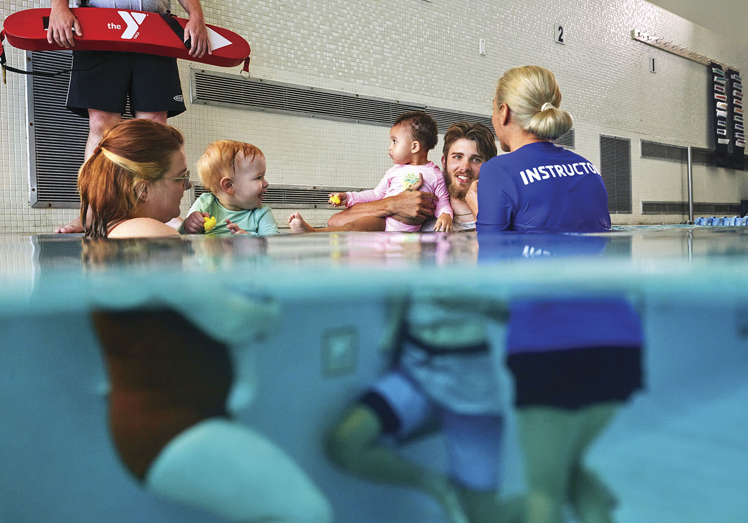 parent-child-banner A parent-and-tot swim class at a pool, showing parents holding babies while an instructor in a blue shirt interacts with them. The photo is partially underwater, highlighting the pool's surface.
