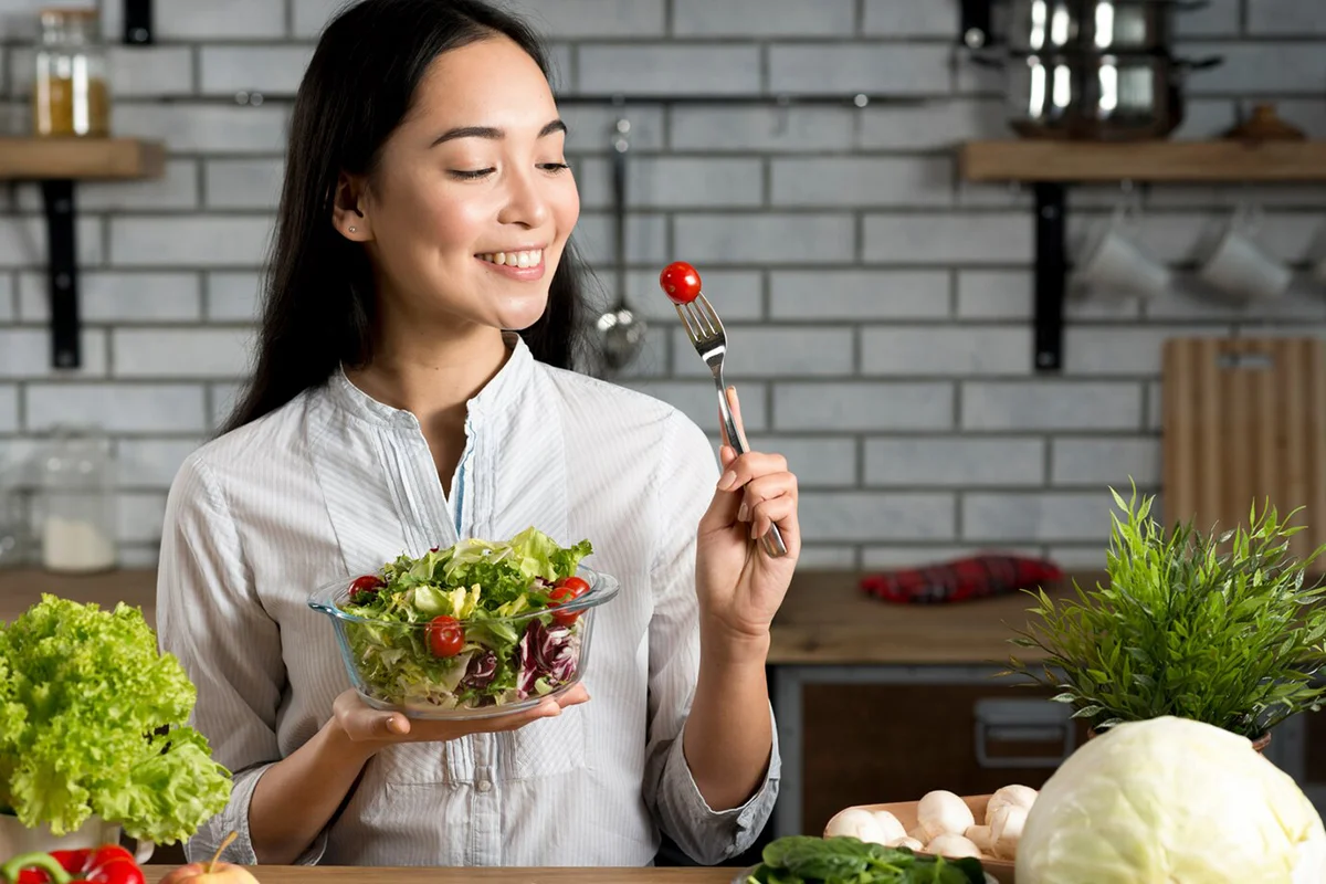 smiling-asian-woman-with-healthy-salad-kitchen_23-2148075983 A woman in a kitchen holding a bowl of fresh salad with lettuce, tomatoes, and greens. She smiles while lifting a cherry tomato on a fork, surrounded by fresh vegetables and herbs on the counter.