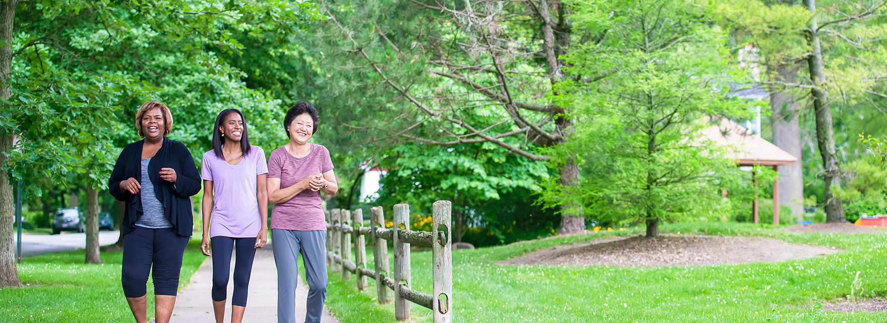 walk-hero Three women enjoy a walk on a shaded path in a green park, surrounded by trees and a wooden fence.