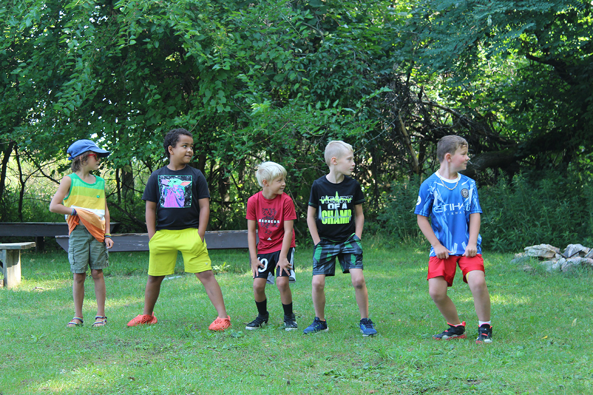 A group of boys stands in a line outdoors, participating in an activity on a grassy field near trees.