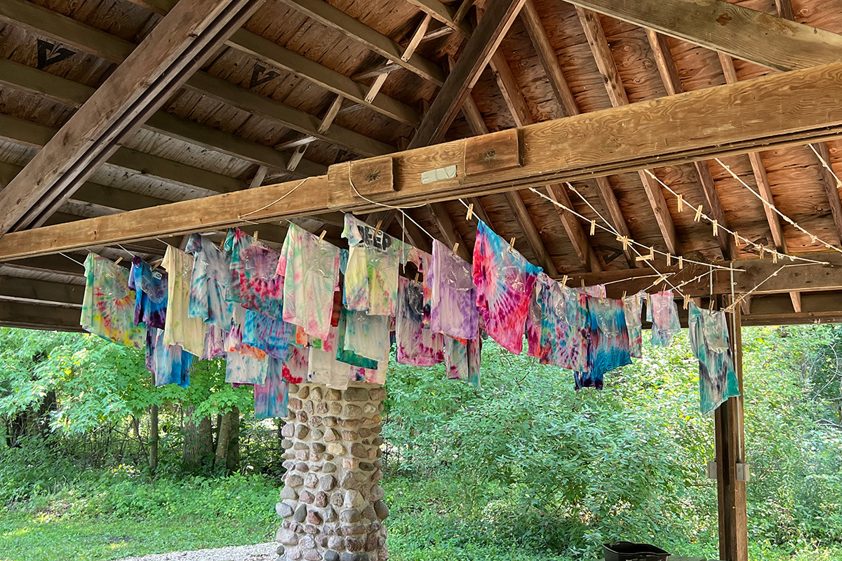 Tie-dye shirts hang on multiple clotheslines under a rustic wooden pavilion, drying in a lush outdoor camp setting.