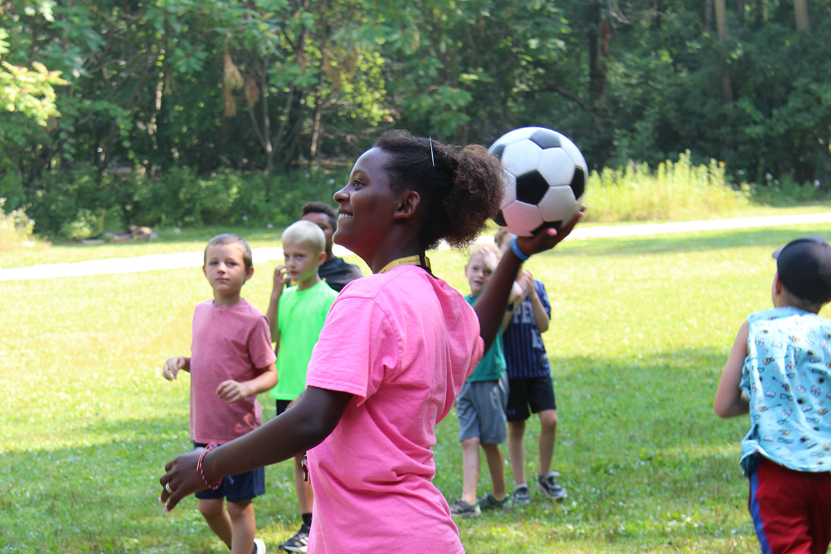 A young girl prepares to throw a soccer ball during an outdoor game with children on a sunny day.