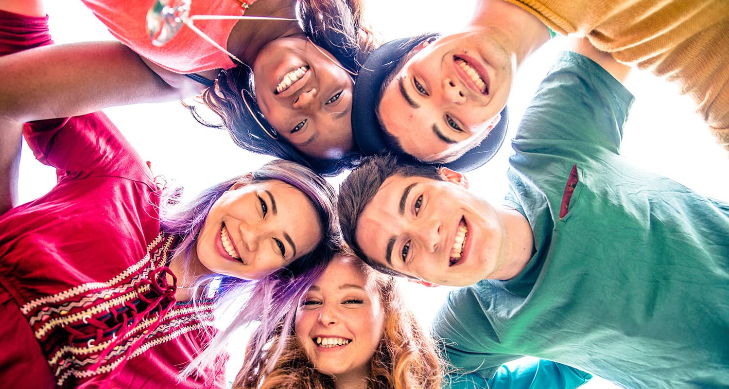 Header_teens-general A group of five smiling friends huddle together, looking down at the camera in a bright, cheerful setting.