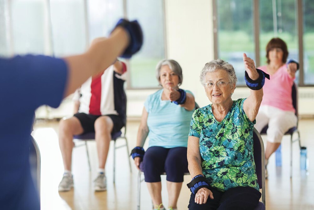 A group of older adults participates in a seated exercise class, using wrist weights to perform arm movements in a bright fitness studio.