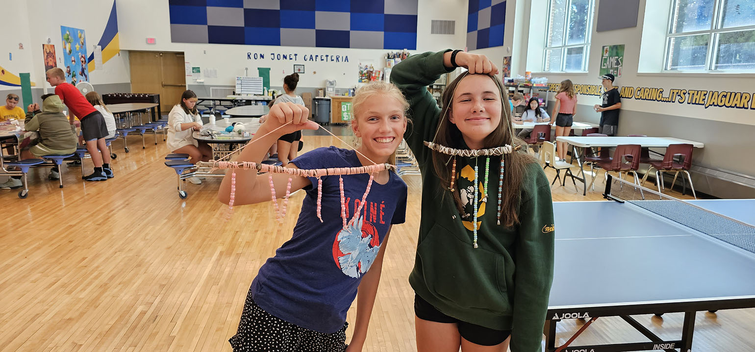 teen-adventure-banner Two girls proudly displaying their handmade bead crafts in a school cafeteria, surrounded by other kids engaged in activities at tables.